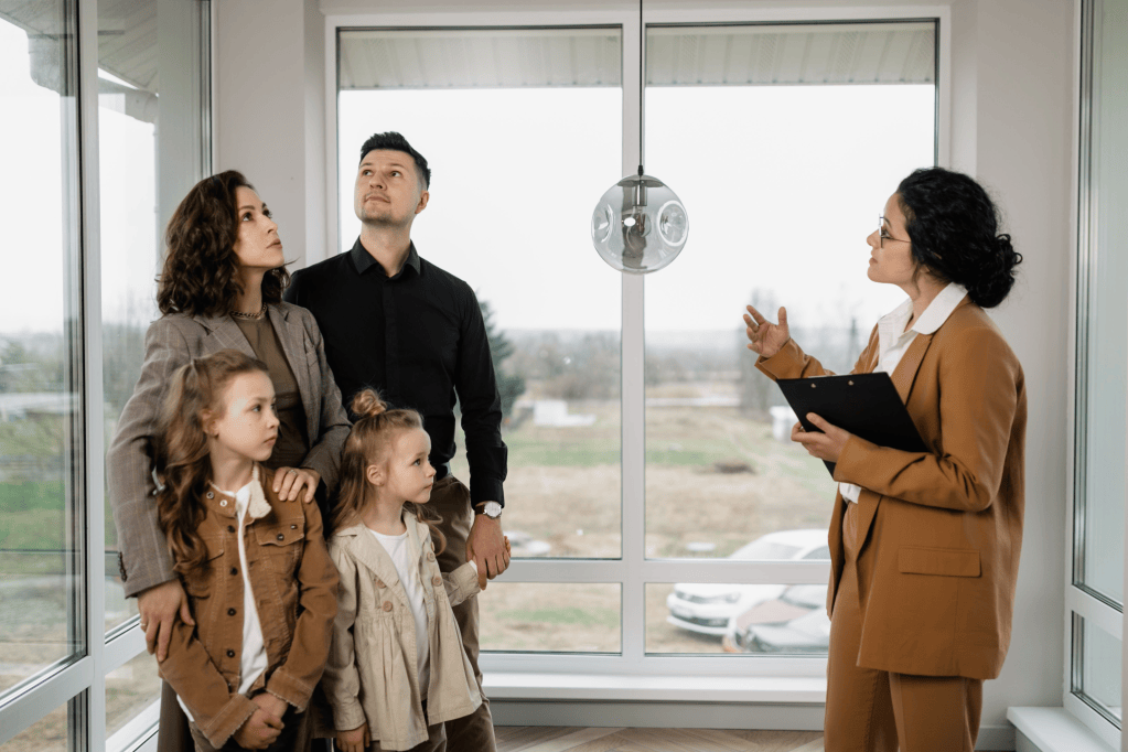 A family of four, including two children, stands indoors looking up at a woman holding a clipboard. The setting features large windows with a view outside, and the woman appears to be explaining something to the family.