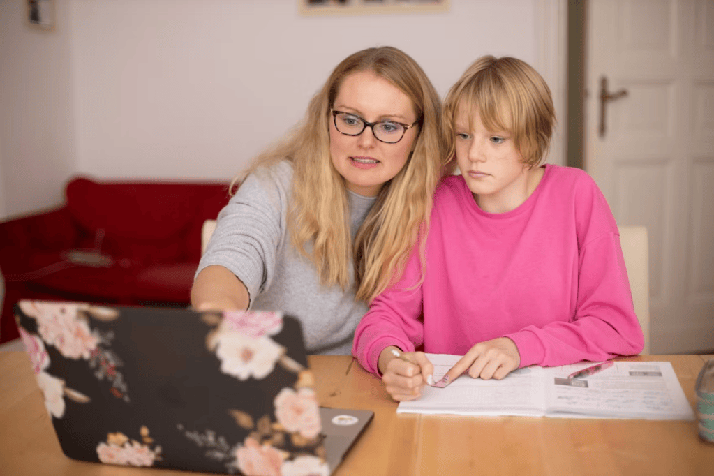 A woman helps a child with schoolwork while sitting at a table with a laptop and notebook.