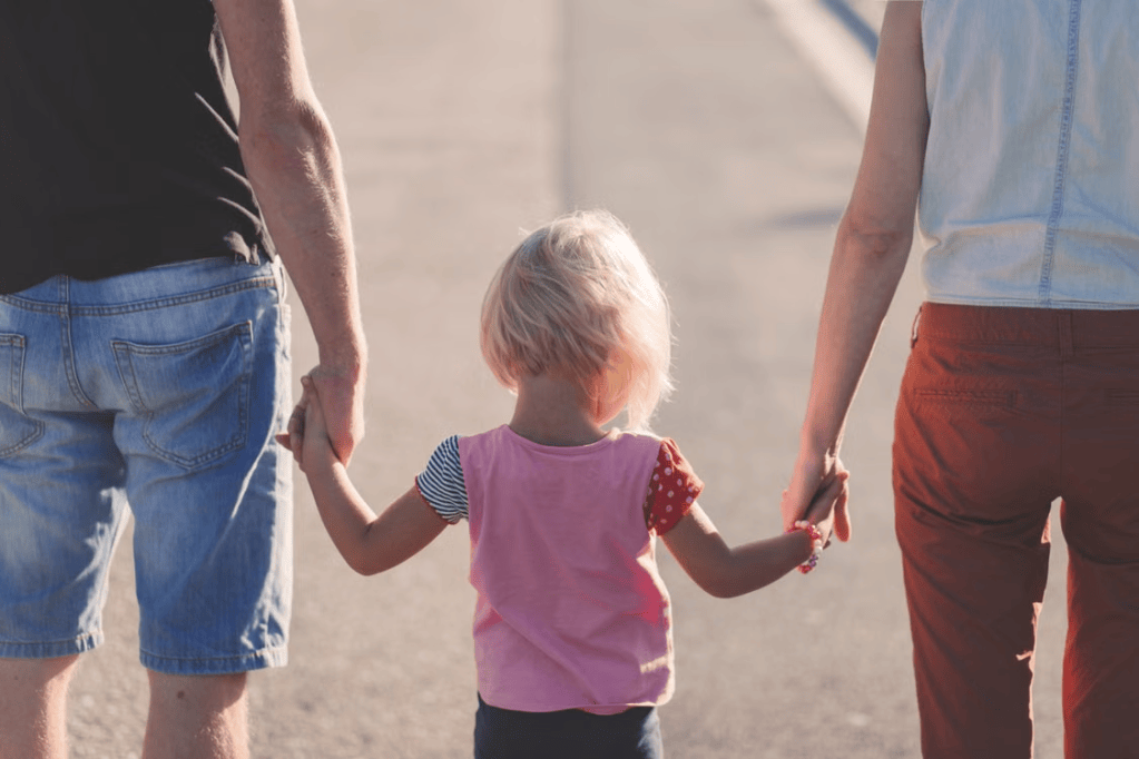 A child holding hands with an adult on either side while walking along a sunlit path.