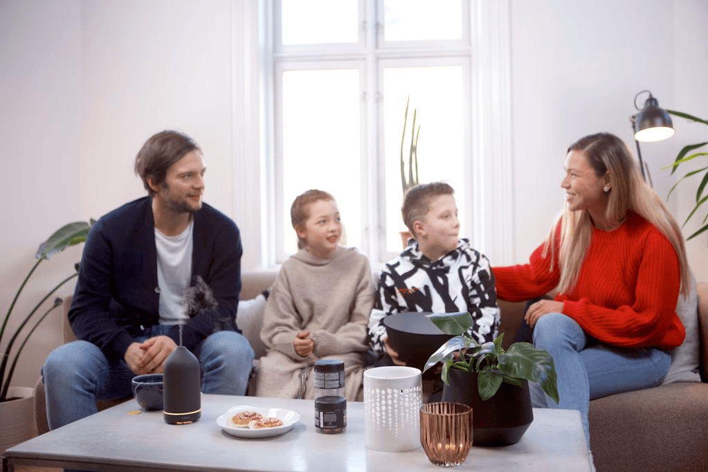 A family of four, including two children, sitting on a cozy sofa in a bright living room. They are engaged in conversation, with a coffee table displaying snacks and plants.