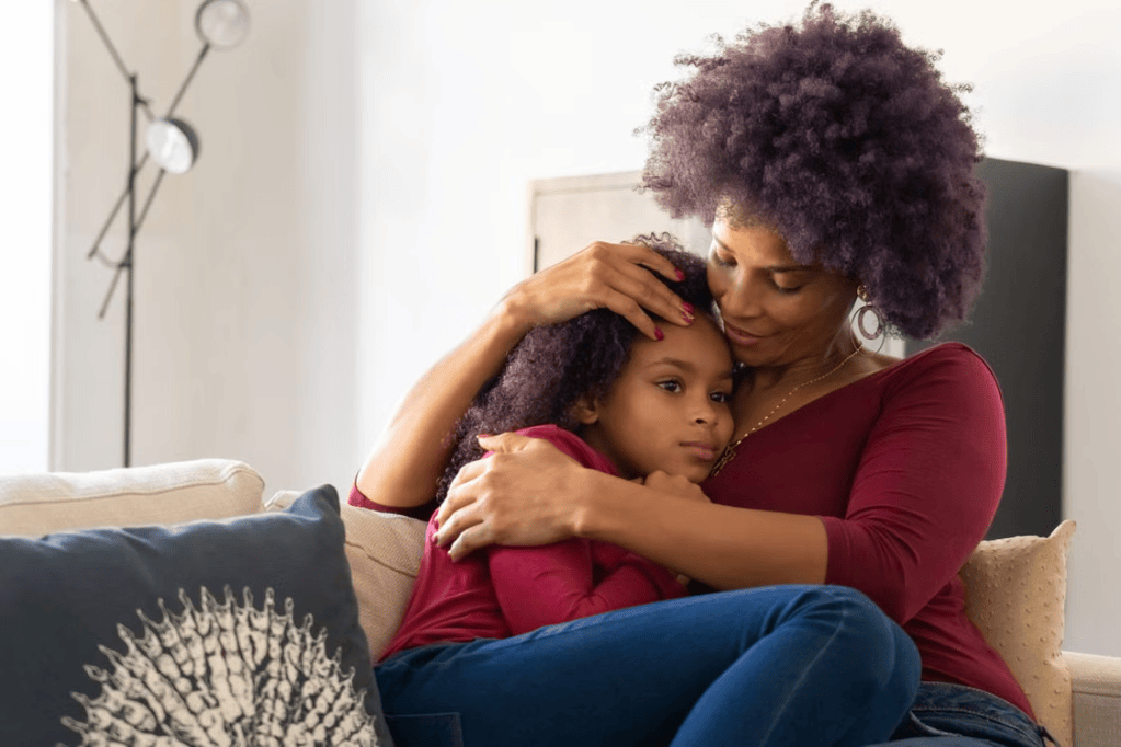 A mother hugs her young daughter on a couch, both wearing matching outfits. The mother shows affection while the child appears to be comforted, in a cozy living room setting.