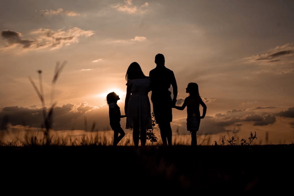 Silhouette of a family of four standing together at sunset, with a warm glow in the background.