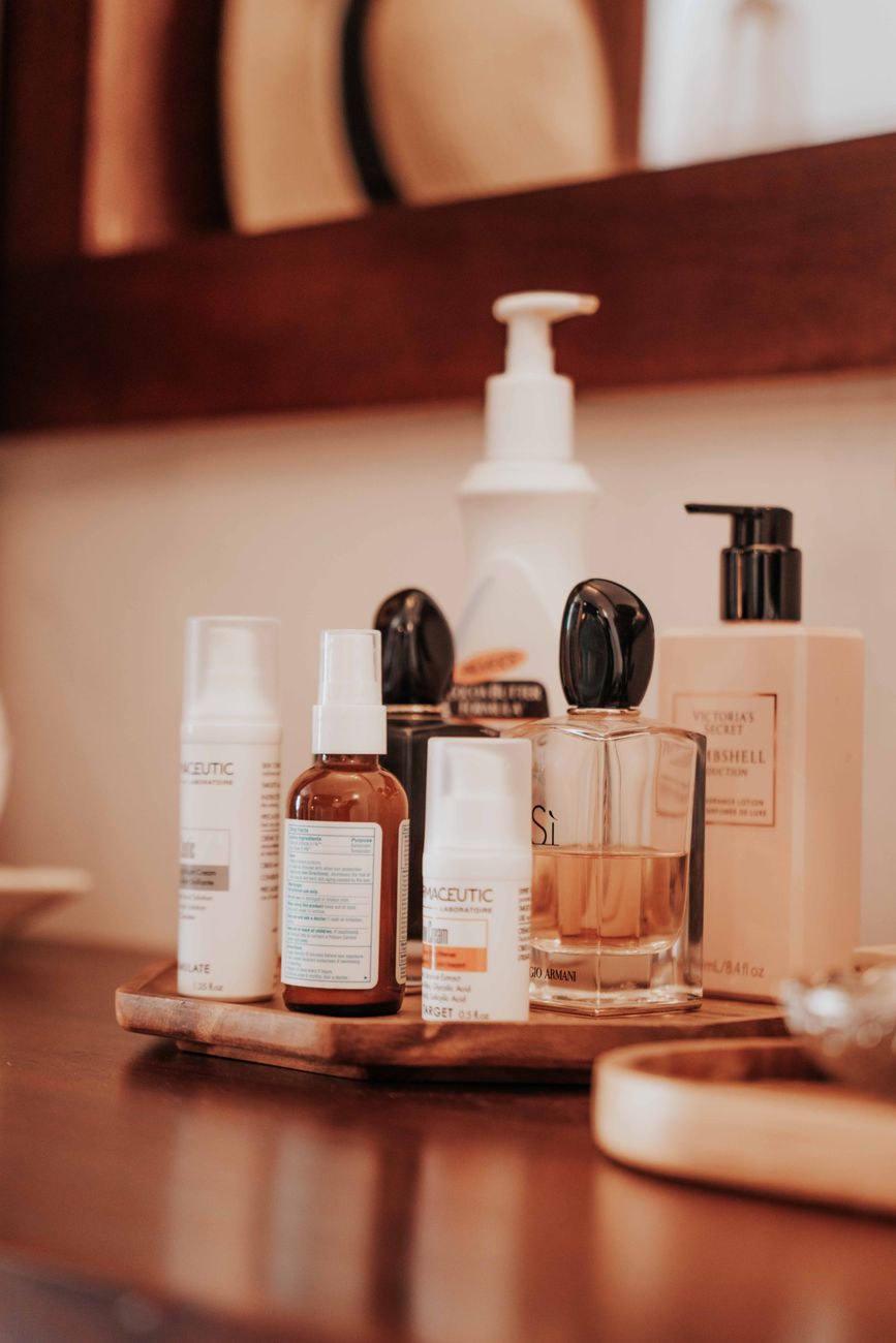 A variety of skincare and fragrance products displayed on a wooden tray, including lotions, serums, and a perfume bottle, set against a mirrored background.