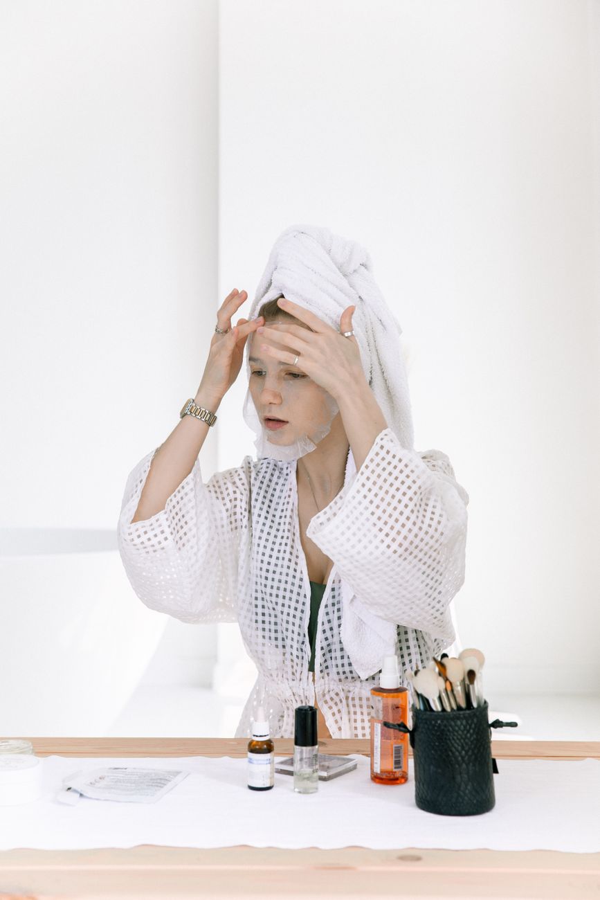 A person applying a face mask while wearing a white robe and towel on their head, with beauty products displayed on a wooden table.