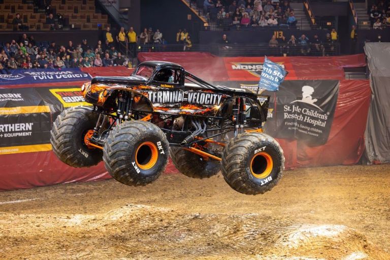 A monster truck named Terminator Velocity performing a jump over a dirt ramp during a live motorsports event, with a crowd in the background.