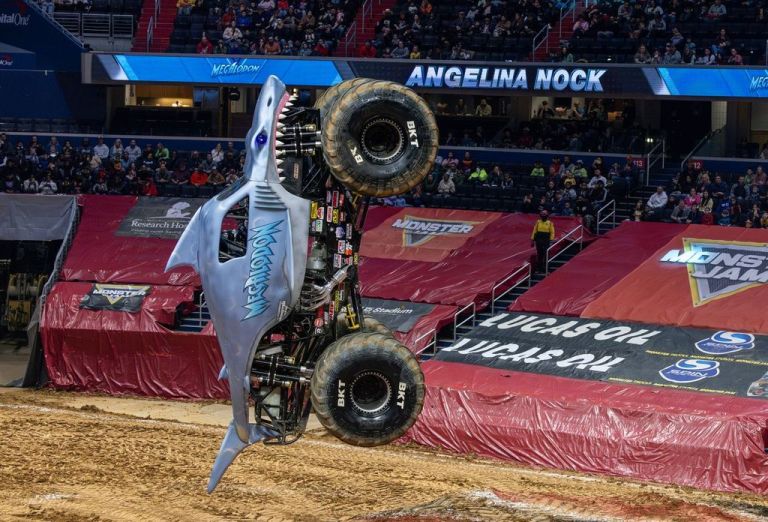 A monster truck named 'Megadon' performs a trick in a stadium arena, showcasing its shark-themed design as it flips upside down during a monster truck event.