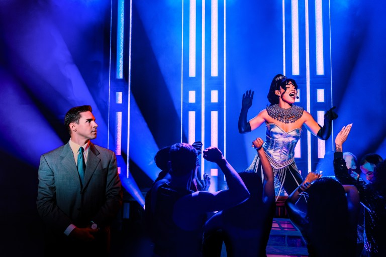 A performer in an elegant outfit captivates the audience with a dynamic pose on stage, illuminated by vibrant blue lights, while a man in a suit watches from the side.