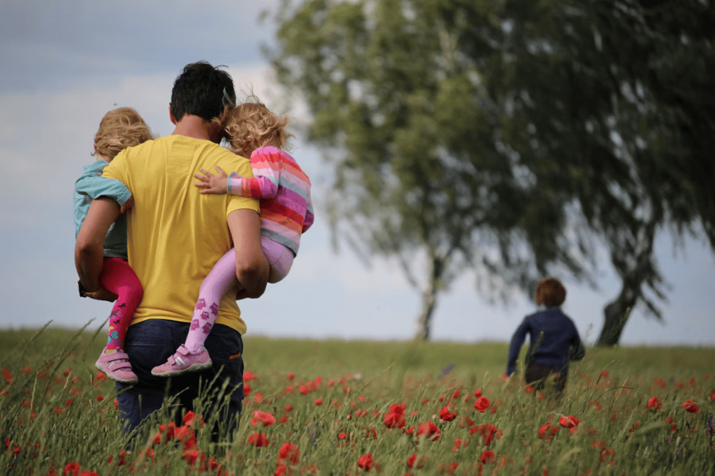 A father carrying two young daughters in a field of red flowers, while a son runs ahead in the background.