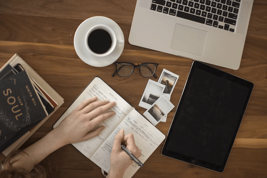 A person's hand writing in a notebook on a wooden table, surrounded by a cup of coffee, eyeglasses, a laptop, an iPad, and several photo prints.