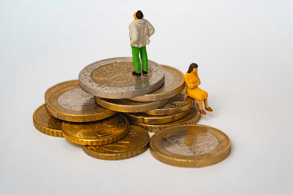 Miniature figures of a man in a white shirt and a woman in a yellow dress standing on a pile of coins, symbolizing finance or investment.