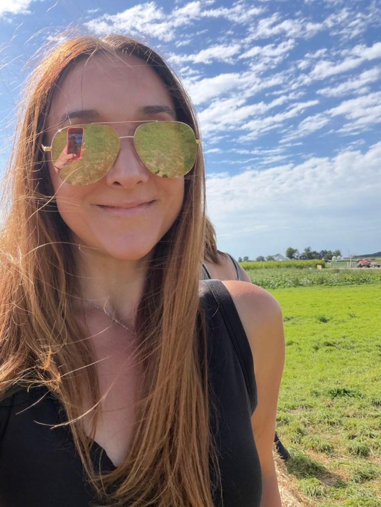 Woman smiling outdoors wearing sunglasses, with green grass and a blue sky in the background.