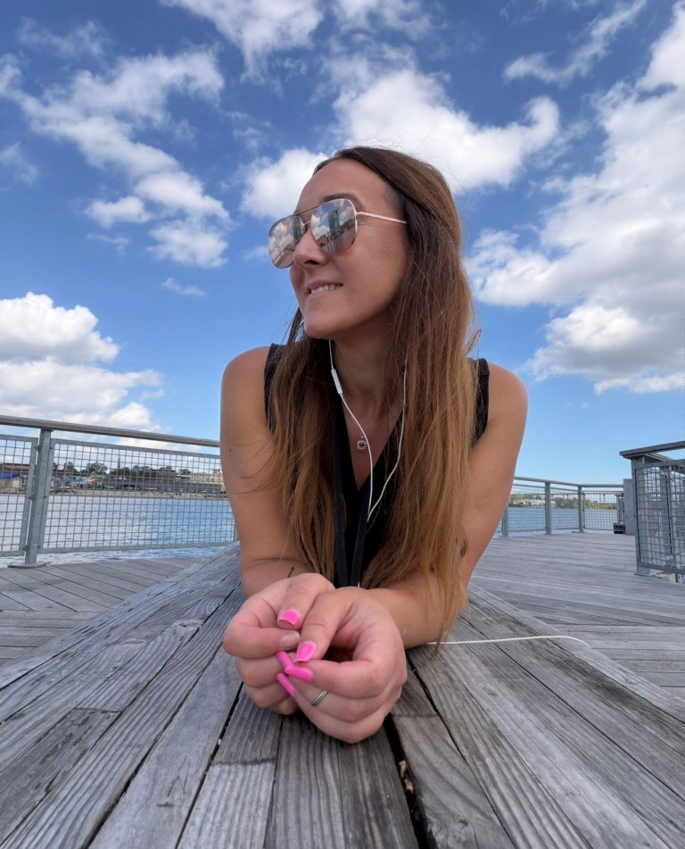 A woman with long brown hair and sunglasses lounging on a wooden deck by the water, listening to music with earphones. The sky is blue with fluffy clouds.