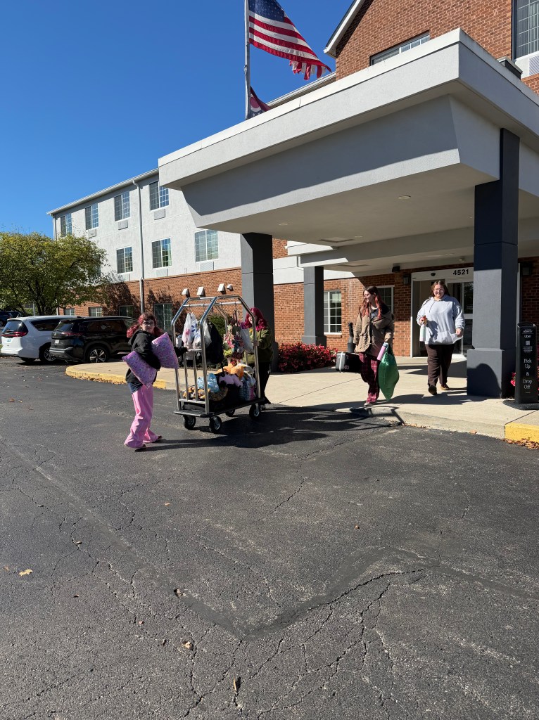 Two women carrying items and a cart with luggage in front of a hotel entrance under a clear blue sky with an American flag.
