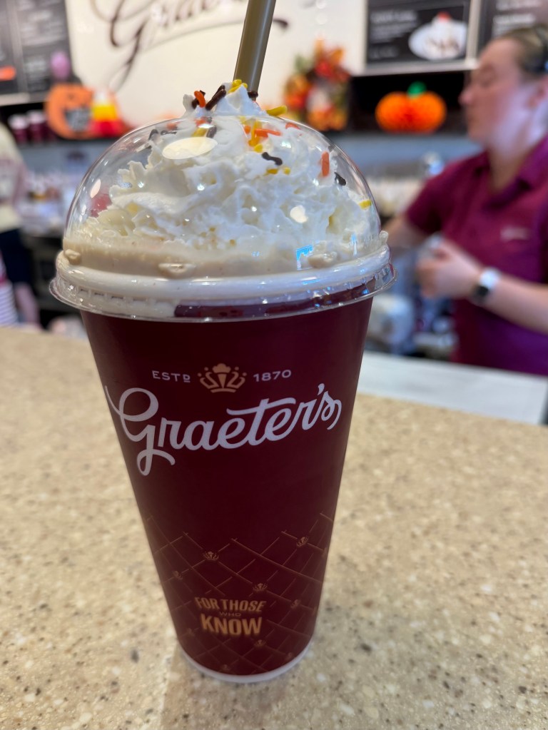 A close-up of a delicious whipped cream topped drink with sprinkles, served in a Graeter's branded cup, placed on a textured countertop. In the blurred background, a staff member is seen working.