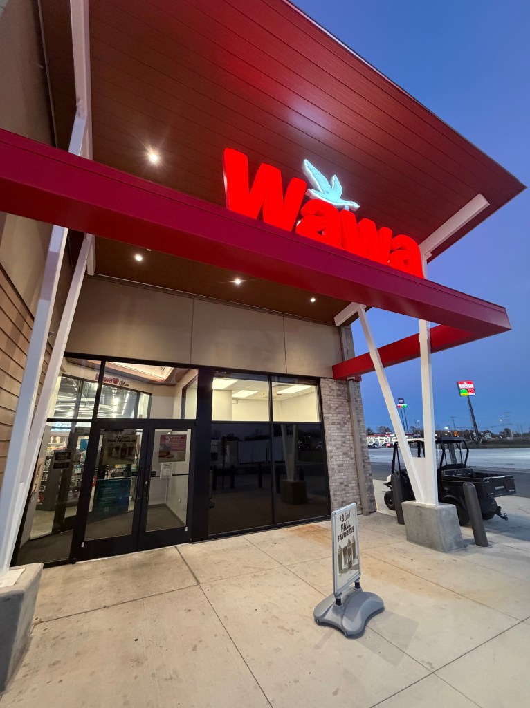 Exterior view of a Wawa convenience store at dusk, featuring a prominent red sign and glass doors, with a sign displayed in front.