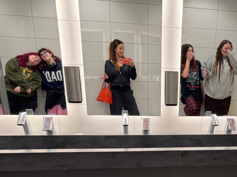 Four women posing in a bathroom mirror, with one taking a selfie while the others lean in playfully.
