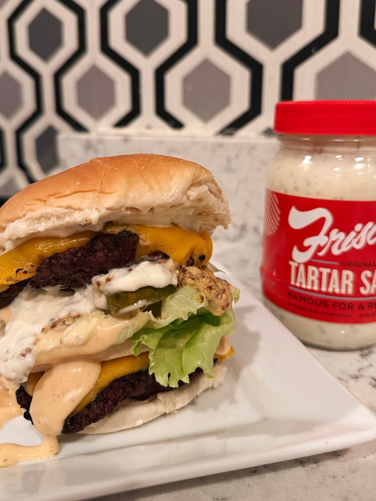 Close-up of a juicy cheeseburger with layers of lettuce, pickles, and sauces, served on a plate beside a jar of tartar sauce.