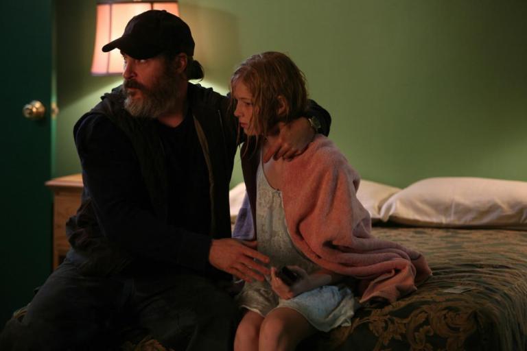 A man with a beard and cap sitting on a bed comforting a young girl wrapped in a pink blanket in a dimly lit room.
