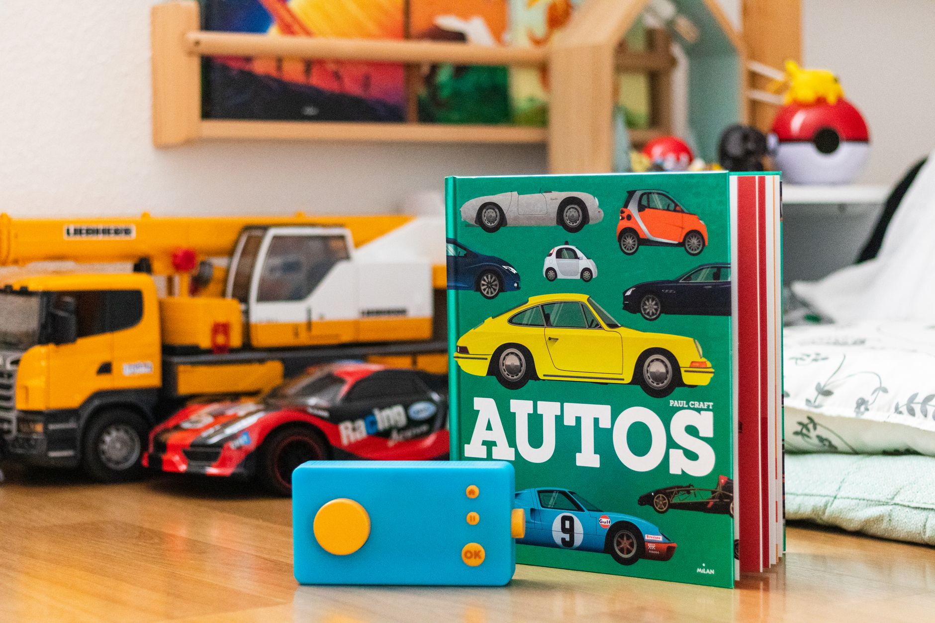 A children's book titled 'AUTOS' featuring various car illustrations, placed next to a blue toy remote and toy cars, including a yellow construction vehicle and a red racing car.