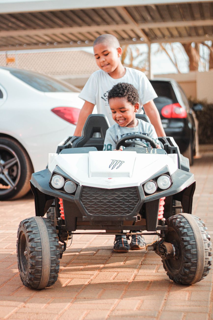 Two boys playing with a small electric ride-on car in a driveway, with one boy steering and the other pushing from behind.