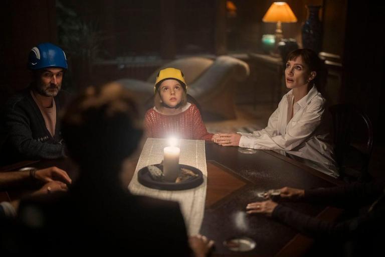 A group of people sitting around a table with a lit candle, featuring a man with a blue helmet, a girl in a yellow helmet, and a woman in a white shirt, in a dimly lit room.
